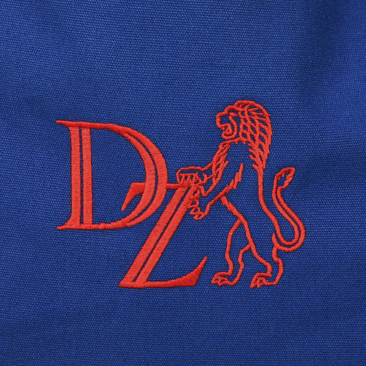 Das Bild zeigt eine hochwertige, blaue Canvas-Tasche mit einer orangefarbenen Stickerei eines Löwen und den Buchstaben 'DZ'.