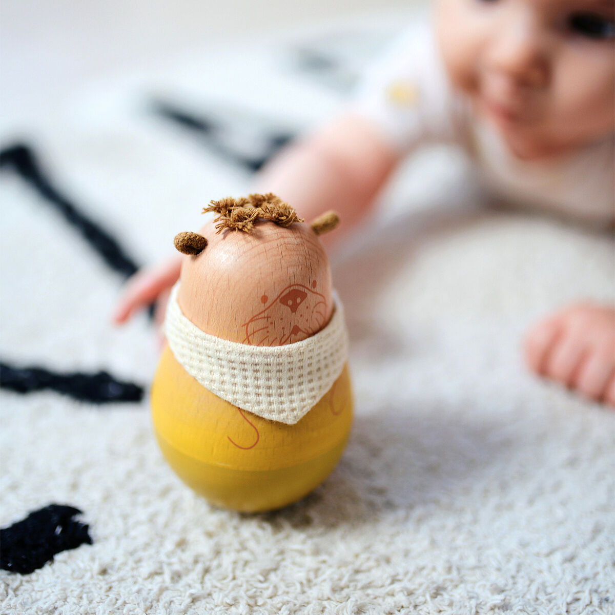 Ein Baby liegt auf einem Teppich und spielt mit einer Stehauffigur aus Holz in gelben und beigen Farben. Das Holzspielzeug hat eine gestrickte Umrandung und eine aufgemalte Verzierung.