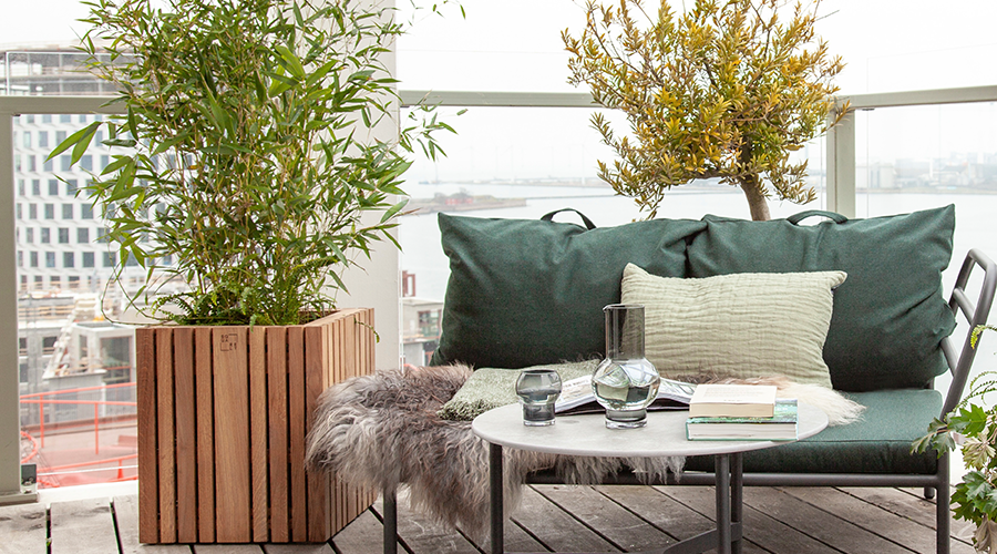 Grüne Sitzbank mit Kissen und Decke auf Holzterrasse, runder Tisch mit Glas und Büchern, große Pflanzen in Holz- und Keramikgefäßen, Stadtblick im Hintergrund