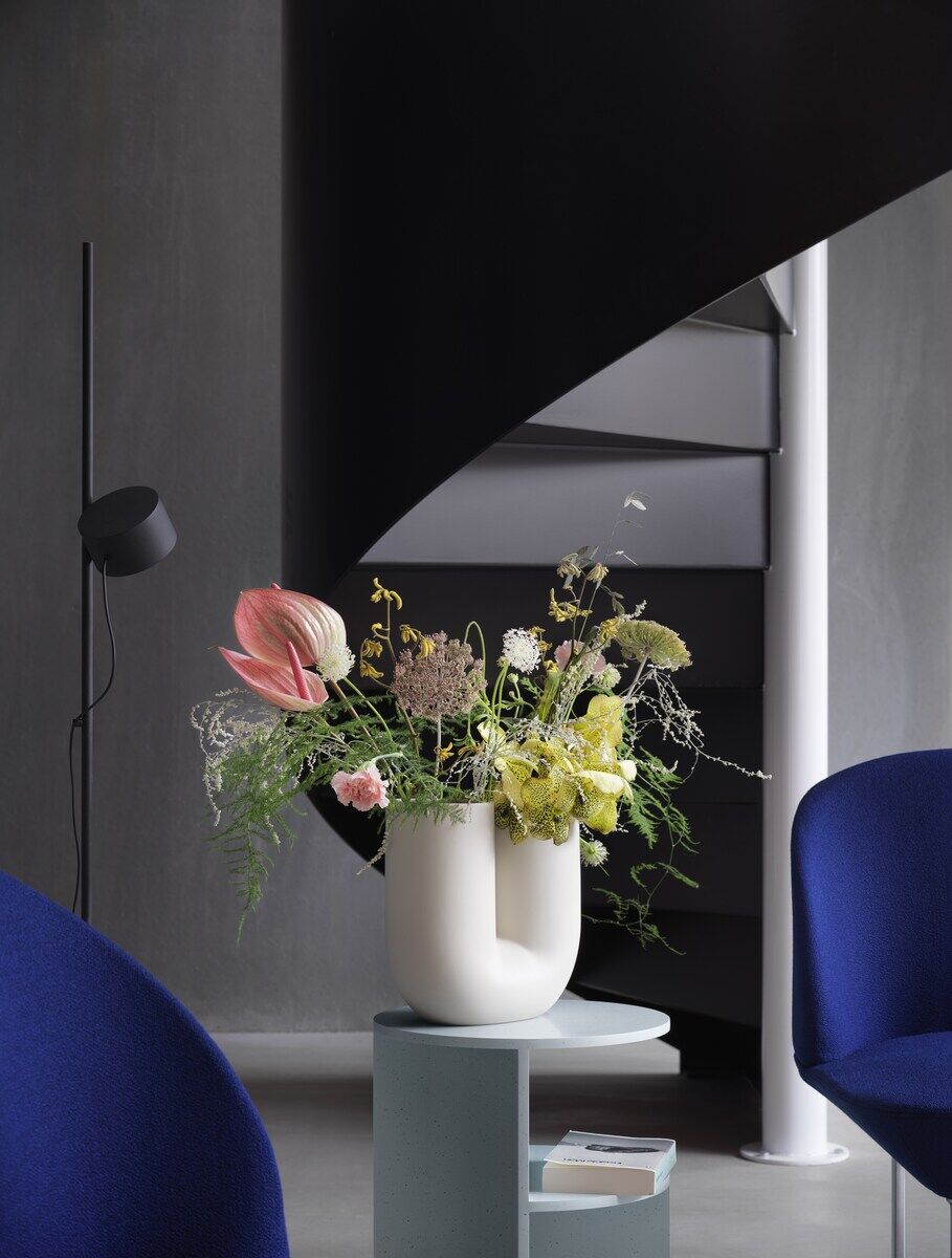 Moderne Bodenlampe in einem stilvollen Wohnzimmer, mit einer schwarzen Struktur. Im Vordergrund steht ein kleiner Tisch mit einer weißen Vase voller bunter Blumen. Im Hintergrund eine Wendeltreppe und zwei blaue Sessel.