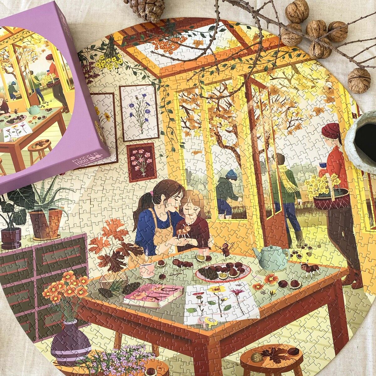 Gezeigtes rundes 1000-Teile-Puzzle »Chestnut Season« von VISSEVASSE zeigt eine herbstliche Innenraumszene mit Familie, Tisch voller Kastanien und Bastelmaterial, warmen Farben und Naturmotiven. Daneben liegt die Schachtel, Deko mit Zweigen, Nüssen und Tannenzapfen.