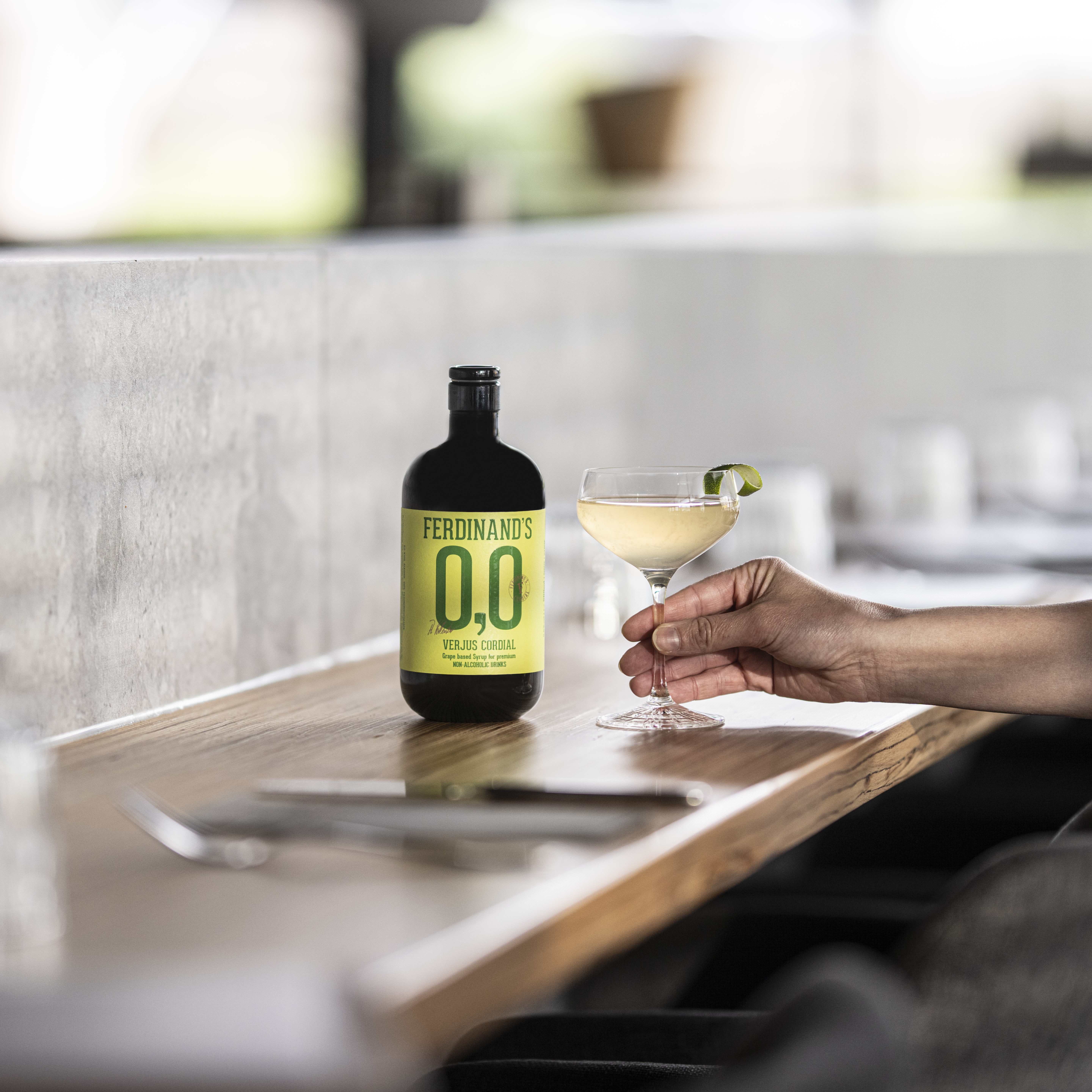 Auf dem Bild steht eine Flasche Ferdinand's Verjus Cordial auf einer hellen Holztheke neben einem Cocktailglas mit einer gelblichen Flüssigkeit. Im Hintergrund ist unscharf eine Fensterfront sowie eine ruhige, moderne Café-Atmosphäre zu erkennen.