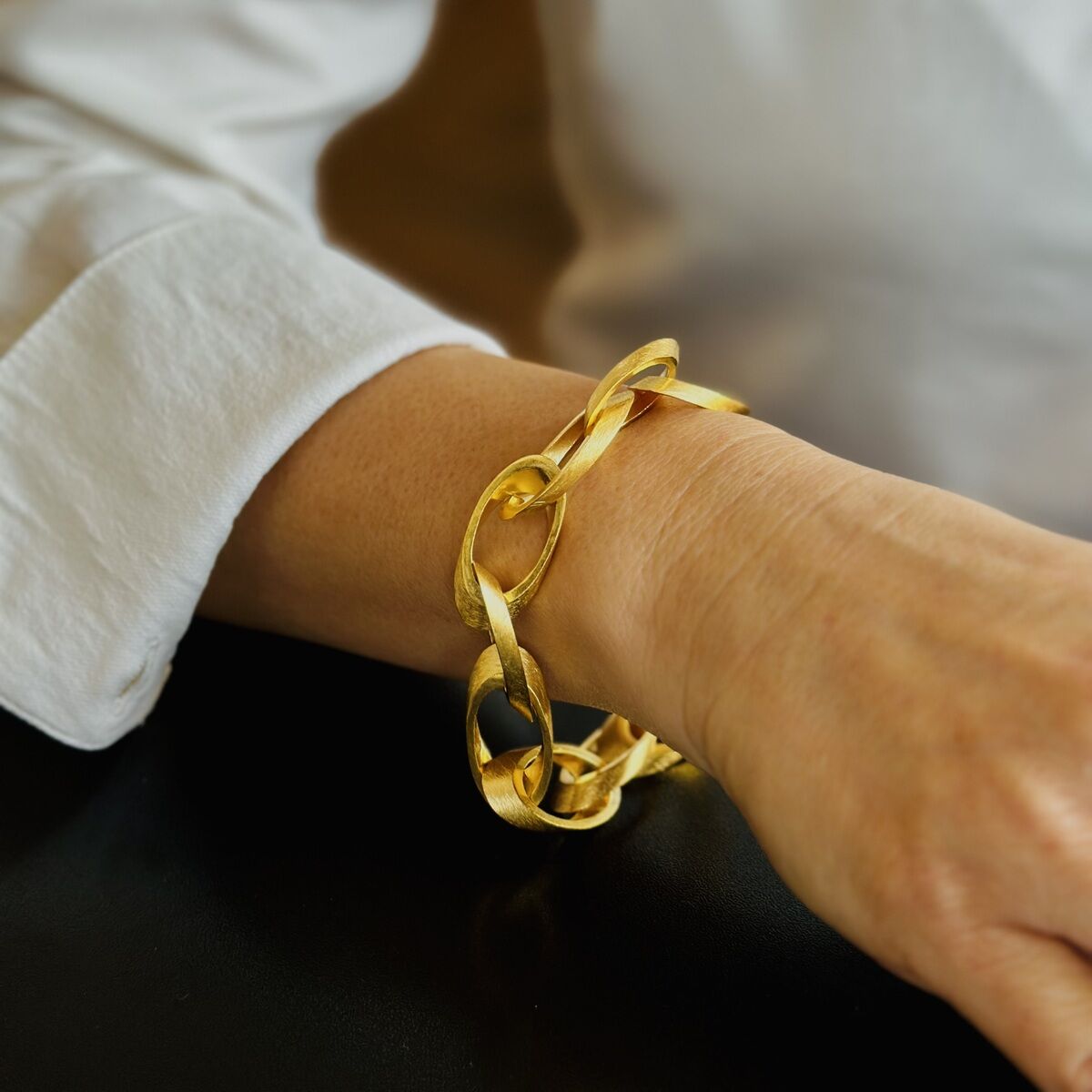 Eine Person trägt ein elegantes Armband aus feingoldplattiertem Silber in Form einer Kette. Der Hintergrund ist verschwommen, die Person trägt ein weißes Oberteil.
