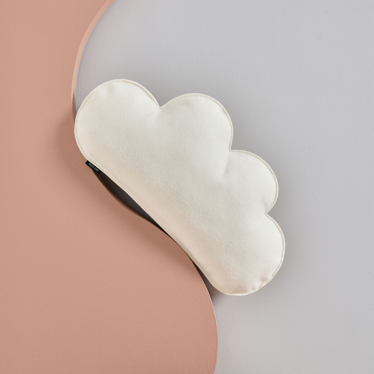 Ein dekoratives Kissen in weißer Wolkenform vor einem Hintergrund mit Pastellrosa und Hellgrau.