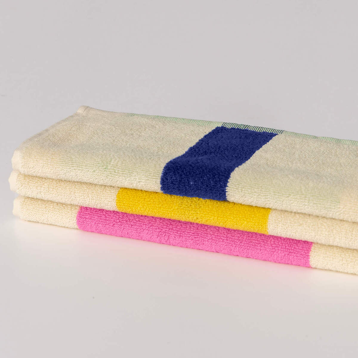 Ein Set aus drei gefalteten Gästehandtüchern liegt auf einem weißen Hintergrund. Die Handtücher sind weiß mit je einem bunten Streifen in Pink, Gelb und Blau in der Mitte.