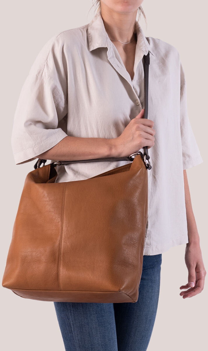 Eine Frau trägt eine braune Beuteltasche »Chaza Pouch Upend« über der rechten Schulter. Sie trägt eine weiße Bluse und blaue Jeans. Die Tasche aus Leder hat einen minimalistischen und eleganten Look.