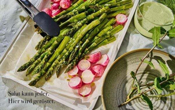 Ofengerichte auf einer weißen Platte: Gebratener grüner Spargel und rosa Radieschen in Scheiben, daneben Metallzange. Daneben ein Teller mit grünem Dip, Kräuterzweig und ein Glas. Sonnenlicht sorgt für kräftige Farben und lebendige Schattierungen.
