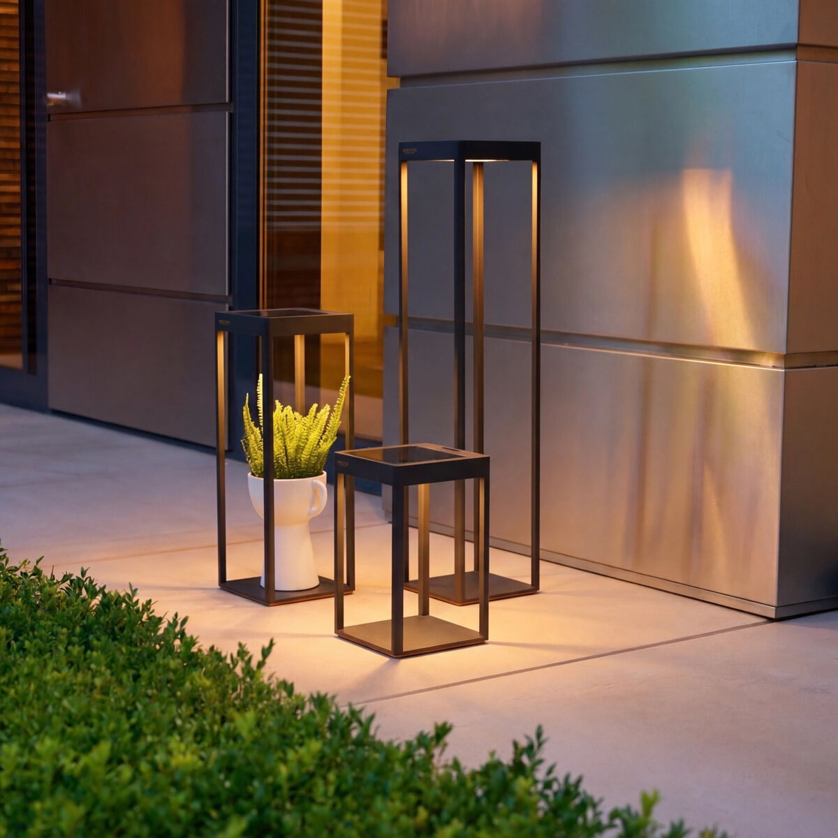 Drei moderne Solarlaternen »WEGA« von Sienagarden aus pulverbeschichtetem Aluminiumdruckguss in Anthrazit stehen auf einer Terrasse neben einer Pflanze. Warmweißes LED-Licht sorgt abends für stimmungsvolle, dekorative Beleuchtung an einer Hauswand aus Metall.