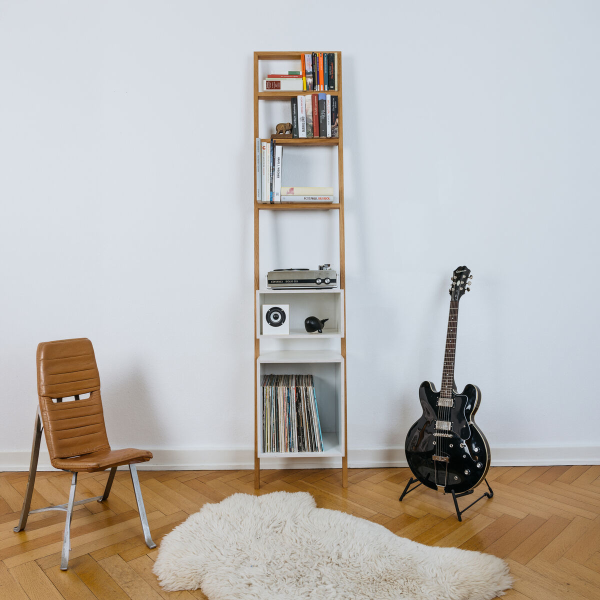 Das Leiterregal »brecht« aus geölter Eiche lehnt an einer weißen Wand. Darauf stehen Bücher, Schallplatten und Musikgeräte. Links befindet sich ein moderner Holzstuhl, rechts lehnt eine schwarze E-Gitarre, davor ein flauschiger, weißer Teppich auf Parkettboden.