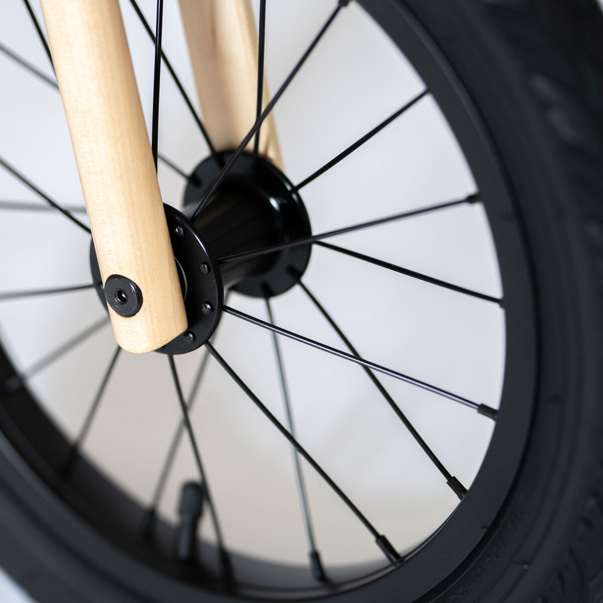 Nahaufnahme eines Vorderrads eines Balance Bike 3in1 mit schwarzer Bereifung und Speichen sowie einer naturfarbenen Holzgabel auf hellem Hintergrund. Das Bild zeigt Details der hochwertigen Verarbeitung und das moderne, kindgerechte Design.