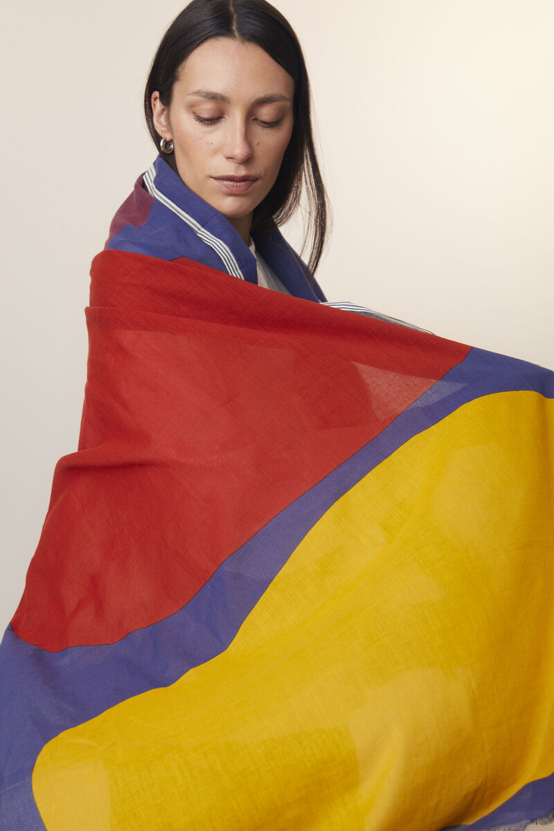 Frau trägt einen Baumwollschal in lebhaften Farben: Rot, Blau und Gelb. Der Schal umhüllt die Schultern locker und verleiht dem Outfit einen stilvollen Akzent.