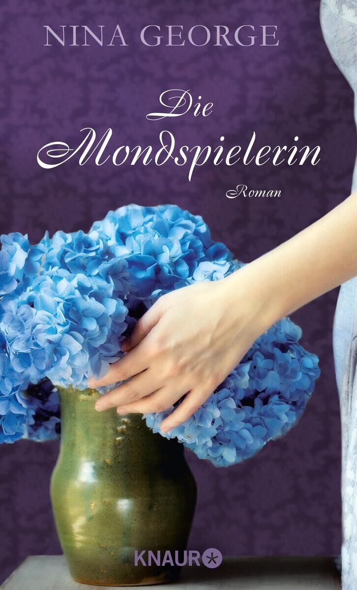 Das Buchcover von 'Die Mondspielerin' von Nina George zeigt eine Vase mit blau-violetten Blüten, eine Hand, die nach den Blüten greift, und einen lila Hintergrund. Oben ist der Name der Autorin und der Titel des Buches in eleganter Schrift dargestellt.