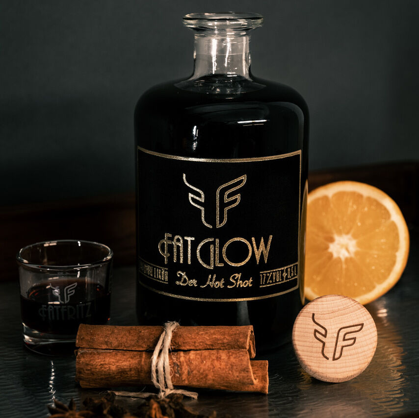 Eine Likörflasche des Glühweinlikörs »FatGlow«, umgeben von einem Shotglas mit Glühweinlikör, einer angeschnittenen Orange, Zimtstangen und Sternanis auf einem Tisch. Die Verpackung der Flasche ist schwarz mit goldenen Akzenten.