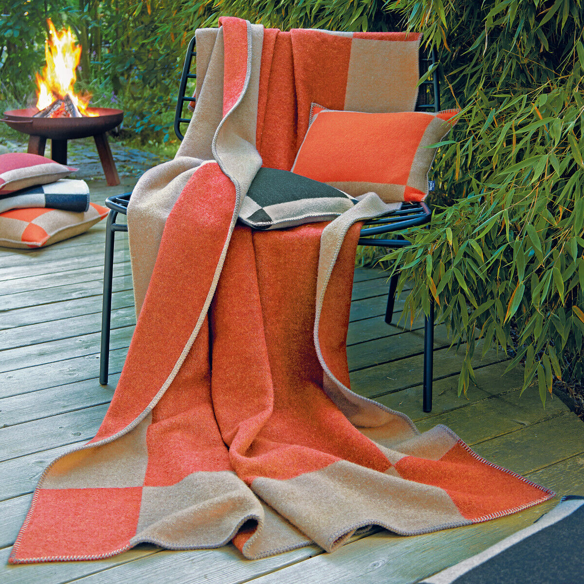 Eine stilvolle Decke mit orange-grauen Quadraten aus Wolle, drapiert über einem Stuhl auf einer Terrasse, flankiert von Kissen und einem Feuerkorb im Hintergrund. Perfekt für gemütliche Abende im Freien.