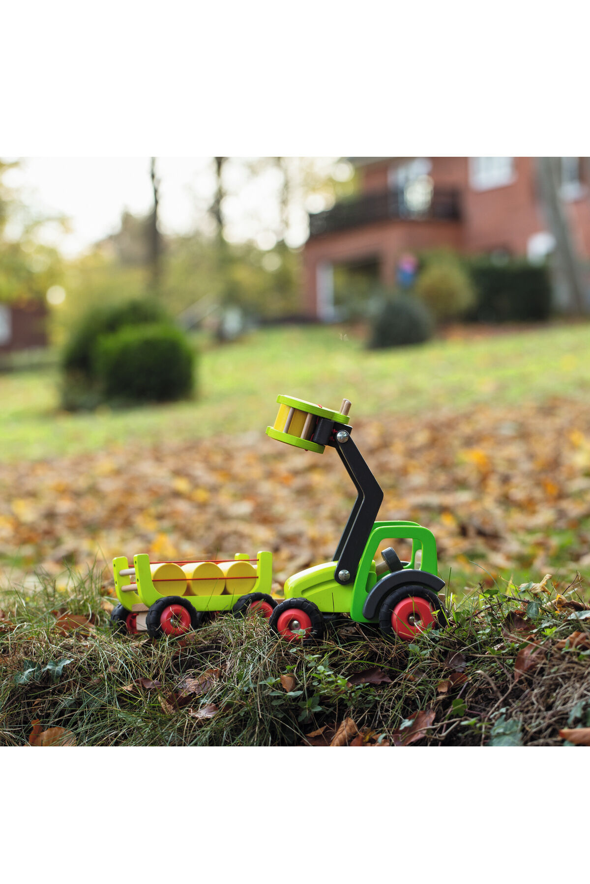 Holzspielzeug-Frontlader mit Heuwagen steht im Freien auf Laub, im Hintergrund ein Haus und grüne Wiese, Herbststimmung, Fokus auf das detailreiche, farbige Spielzeugtraktor-Set.