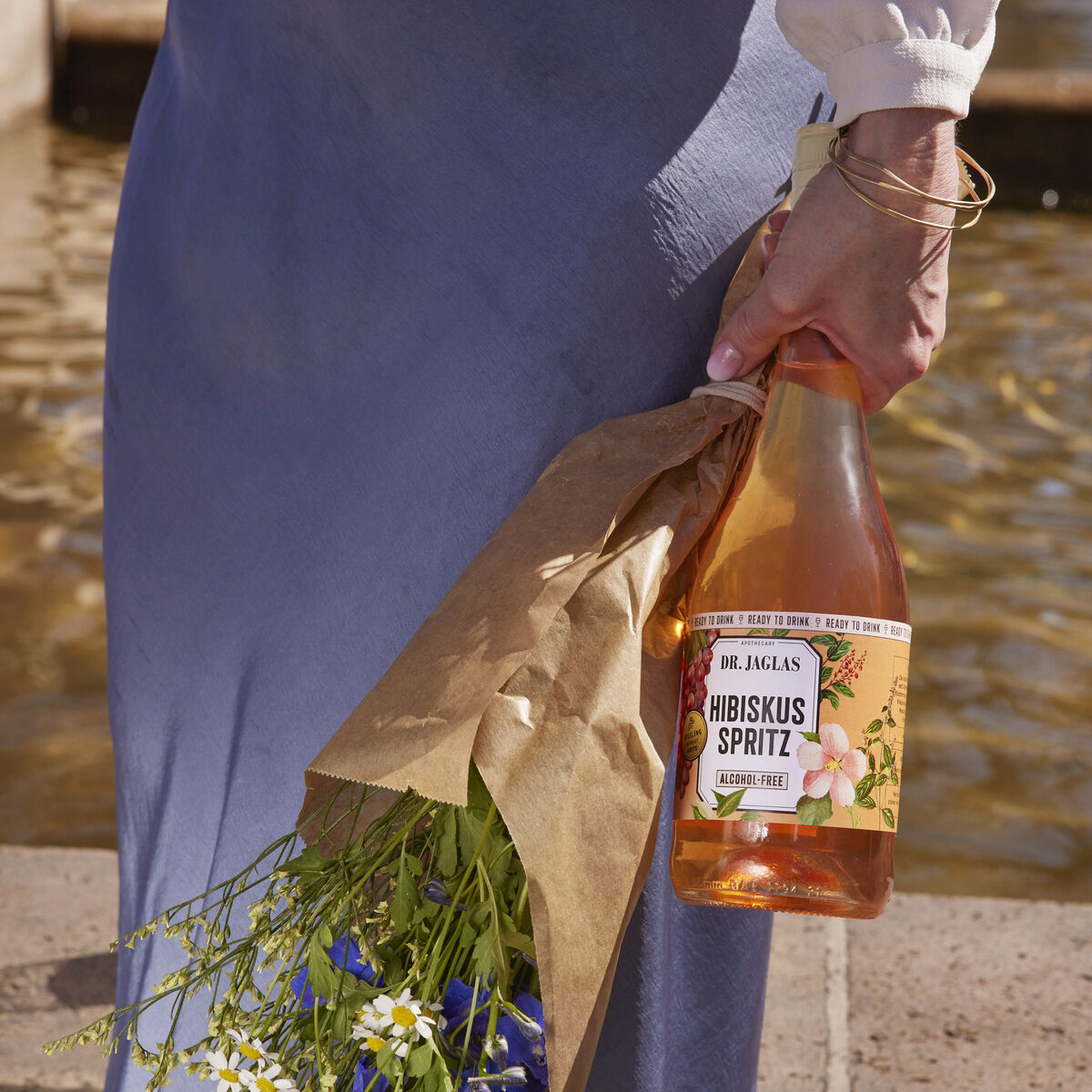 Eine Person in einem blauen Kleid hält einen Blumenstrauß und eine Flasche Hibiskus Spritz. Die Flasche ist in Packpapier eingewickelt und es sind blaue und weiße Blumen sichtbar. Im Hintergrund befindet sich ein Wasserbrunnen.
