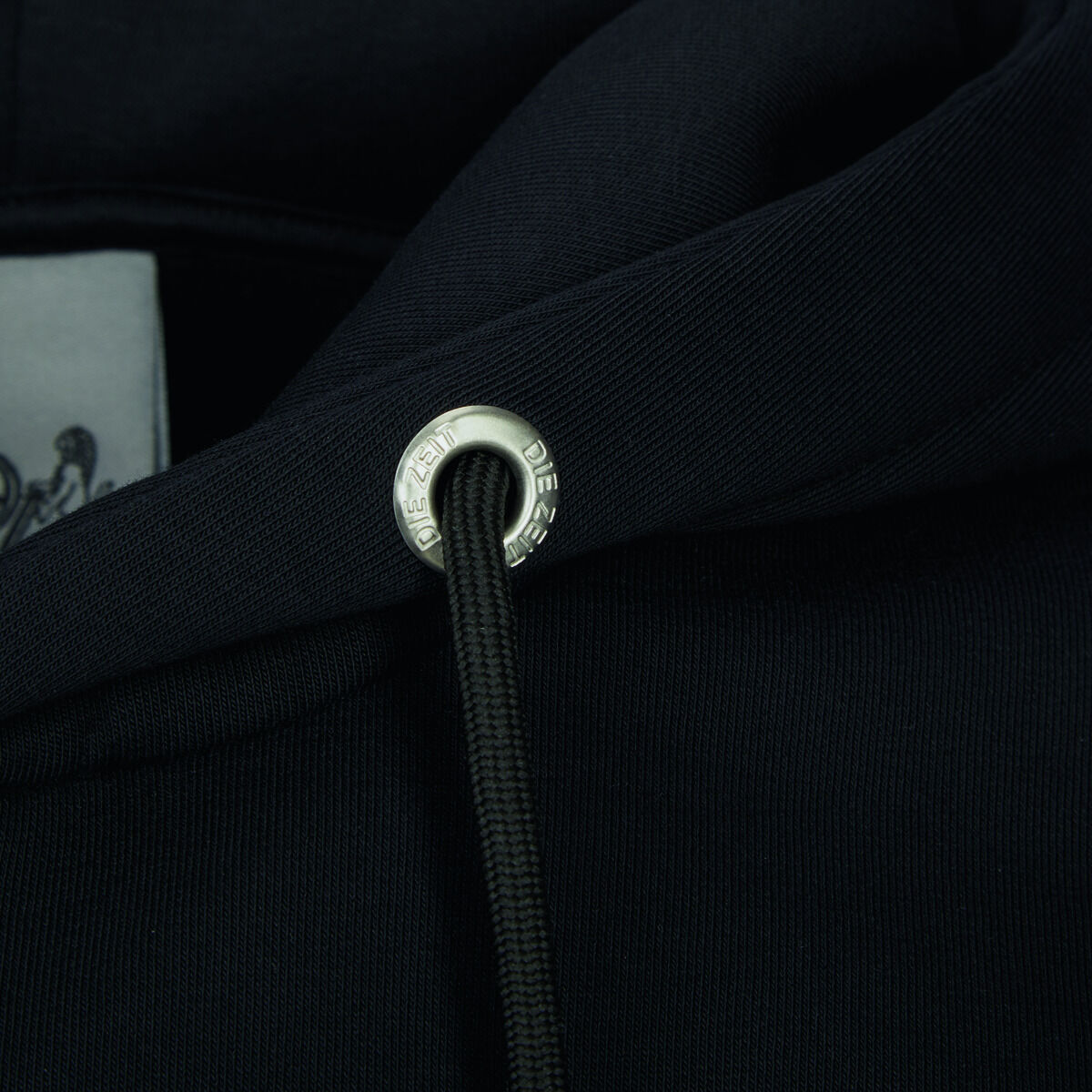 Nahaufnahme eines schwarzen Hoodies aus der ZEIT-Kollektion mit einem edlen Kordelstopper und Detailansicht des Markenlogos.