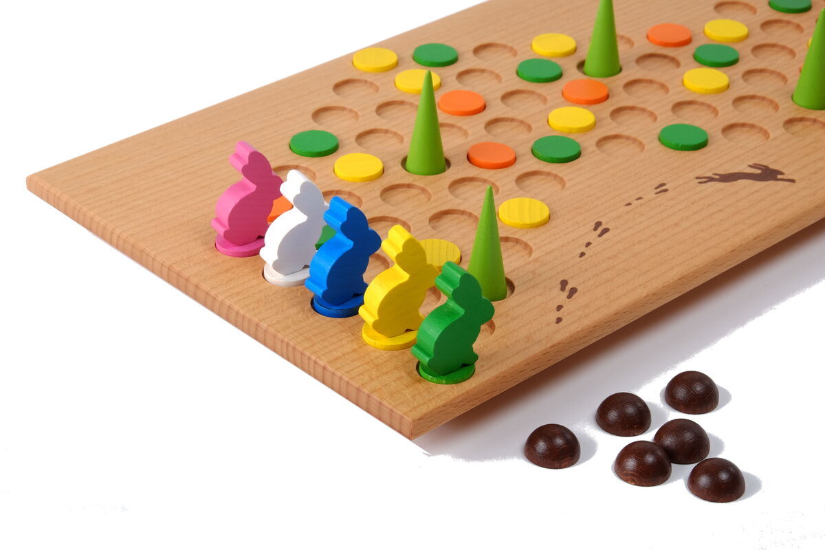 Brettspiel namens 'Hakenschlagen' aus Holz, mit bunten, kuppelförmigen Figuren in Farben wie Grün, Gelb, Rot, Blau, Weiß und Pink. Im Vordergrund befinden sich einige Schokoladenstückchen. Das Spielbrett hat mehrere Löcher und grüne Kegel.