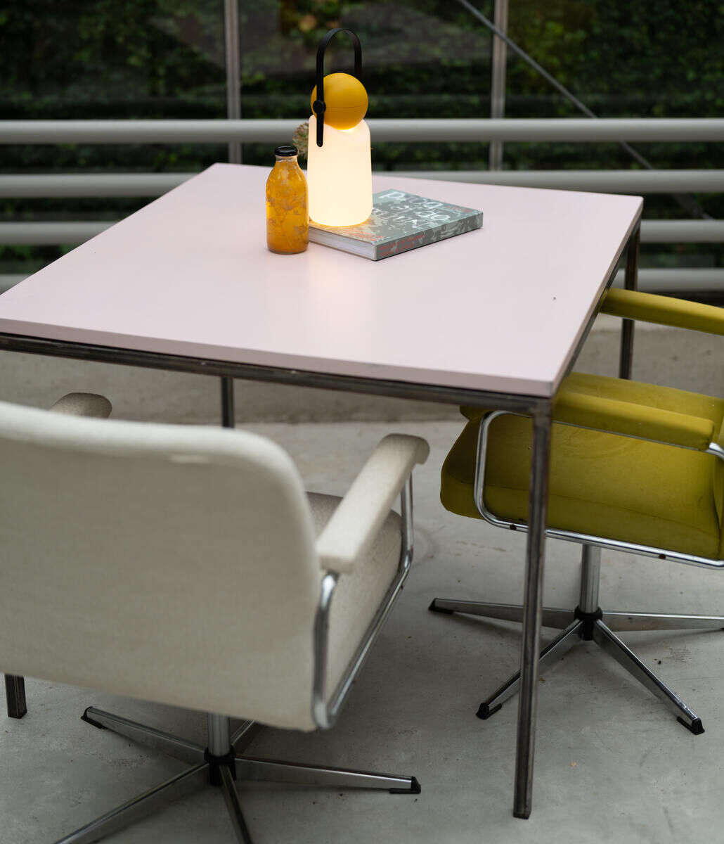 Eine moderne Guidelight Lampe auf einem Tisch mit einem Buch und einer Flasche, umgeben von zwei Stühlen, in einem minimalistischen und gemütlichen Innenraum.