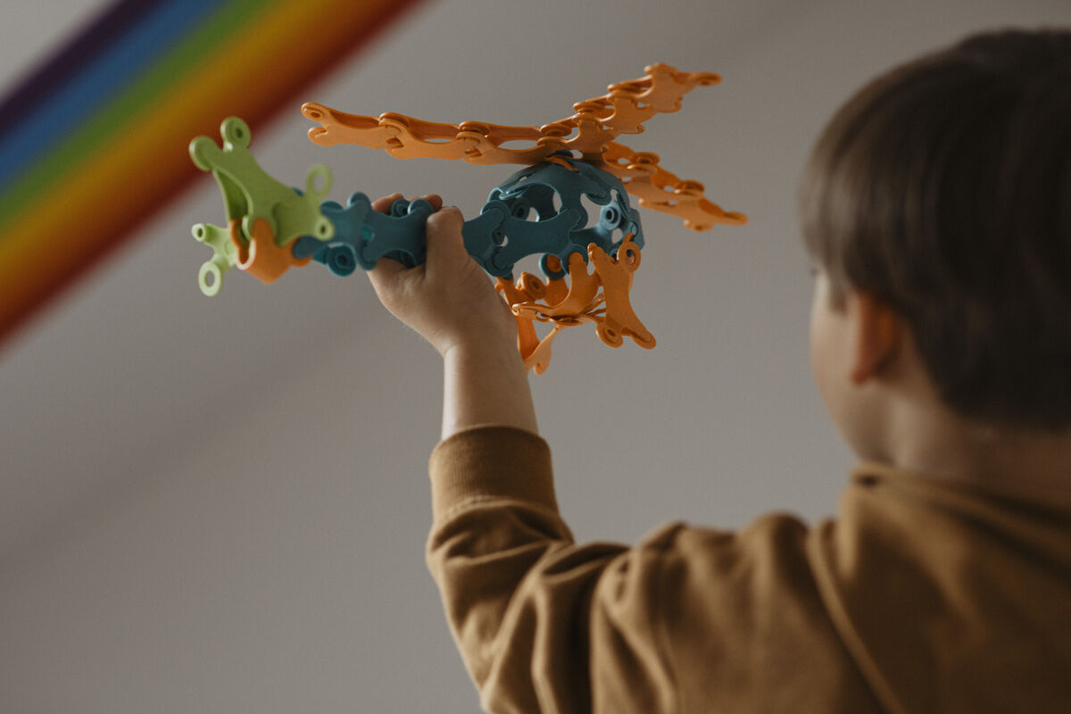 Ein Kind hält ein kreatives Spielzeug aus Binabo-Bausteinen, das in Form eines Drachens zusammengesetzt ist. Im Hintergrund ist ein Regenbogen zu sehen.