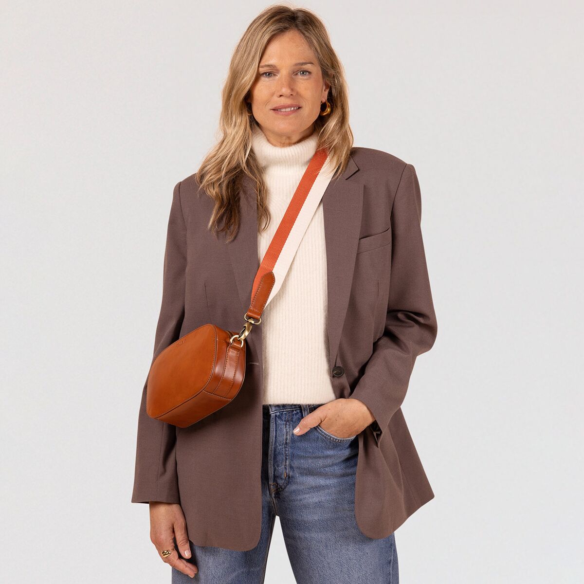 Eine junge Frau steht vor hellem Hintergrund und trägt eine braune Umhängetasche »Maya« aus Leder, kombiniert mit einem cremefarbenen Rollkragenpullover und einem braunen Blazer. Die Tasche ist modern, minimalistisch und wird lässig über die Schulter getragen.