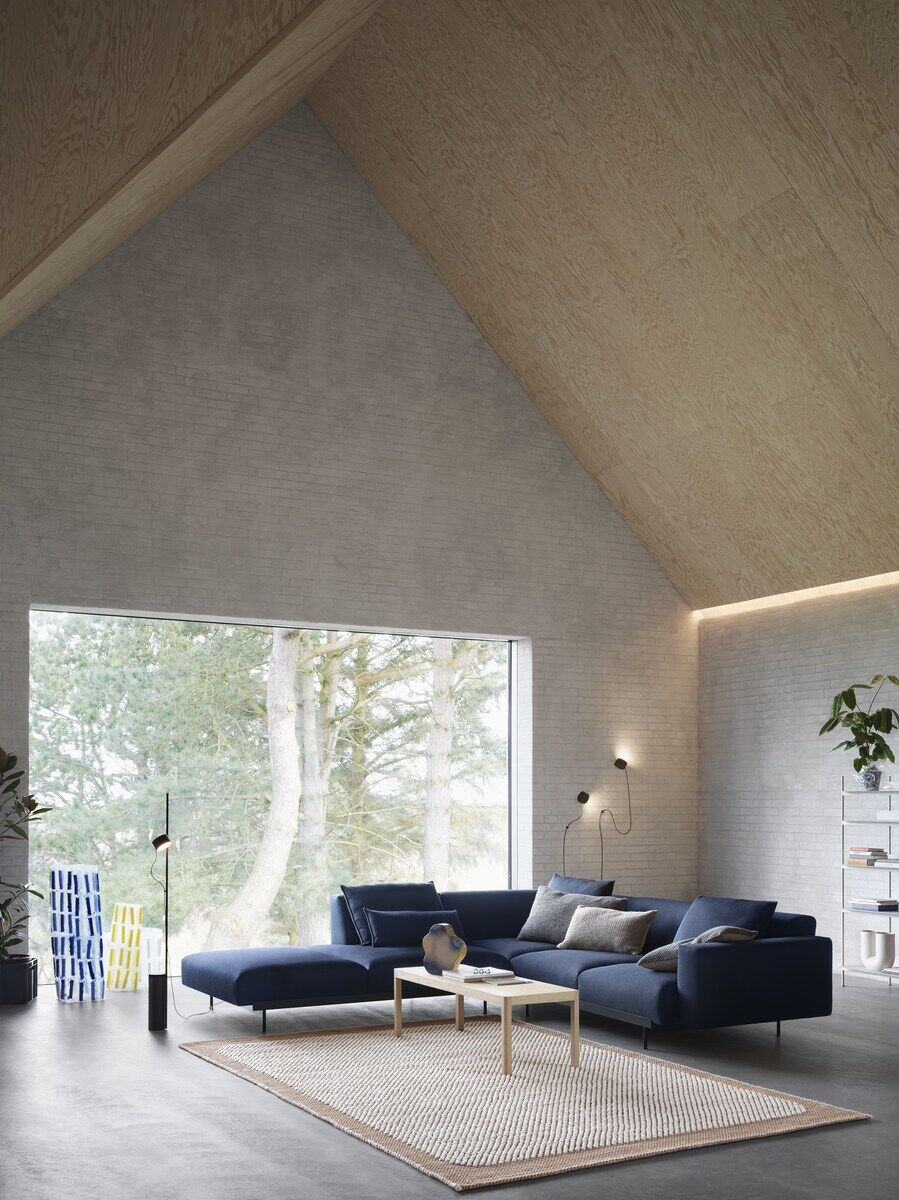 Modernes Wohnzimmer mit großer Fensterfront, blauen Sofa, hellem Holzboden und minimalistischer Bodenlampe »Post«. Die Lampe ist schwarz und steht neben einem Beistelltisch aus Holz. Hinten sind eine Pflanze und ein Regal aus Metall zu sehen.