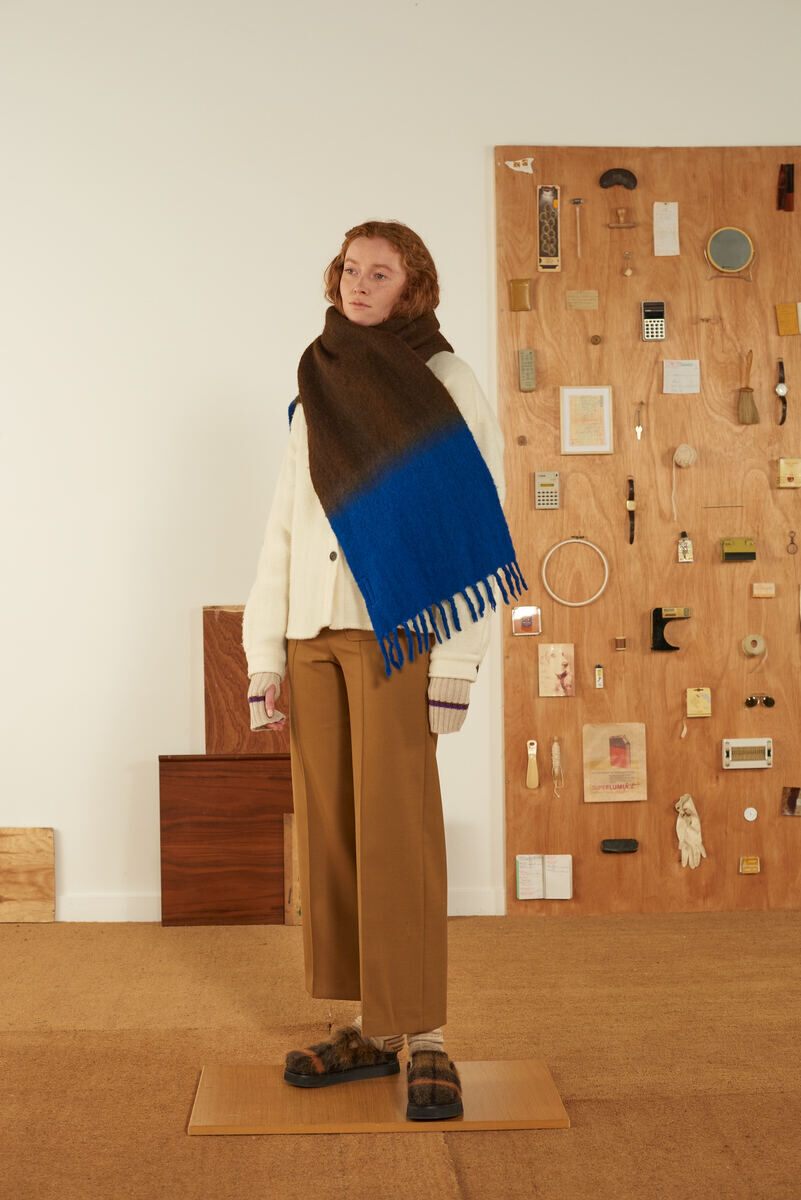 Eine Frau trägt einen großen Schal mit Farbverlauf von Braun zu Blau sowie eine helle Strickjacke und braune Hose. Sie steht in einem Raum mit Holzelementen an der Wand und einem Holzboden.