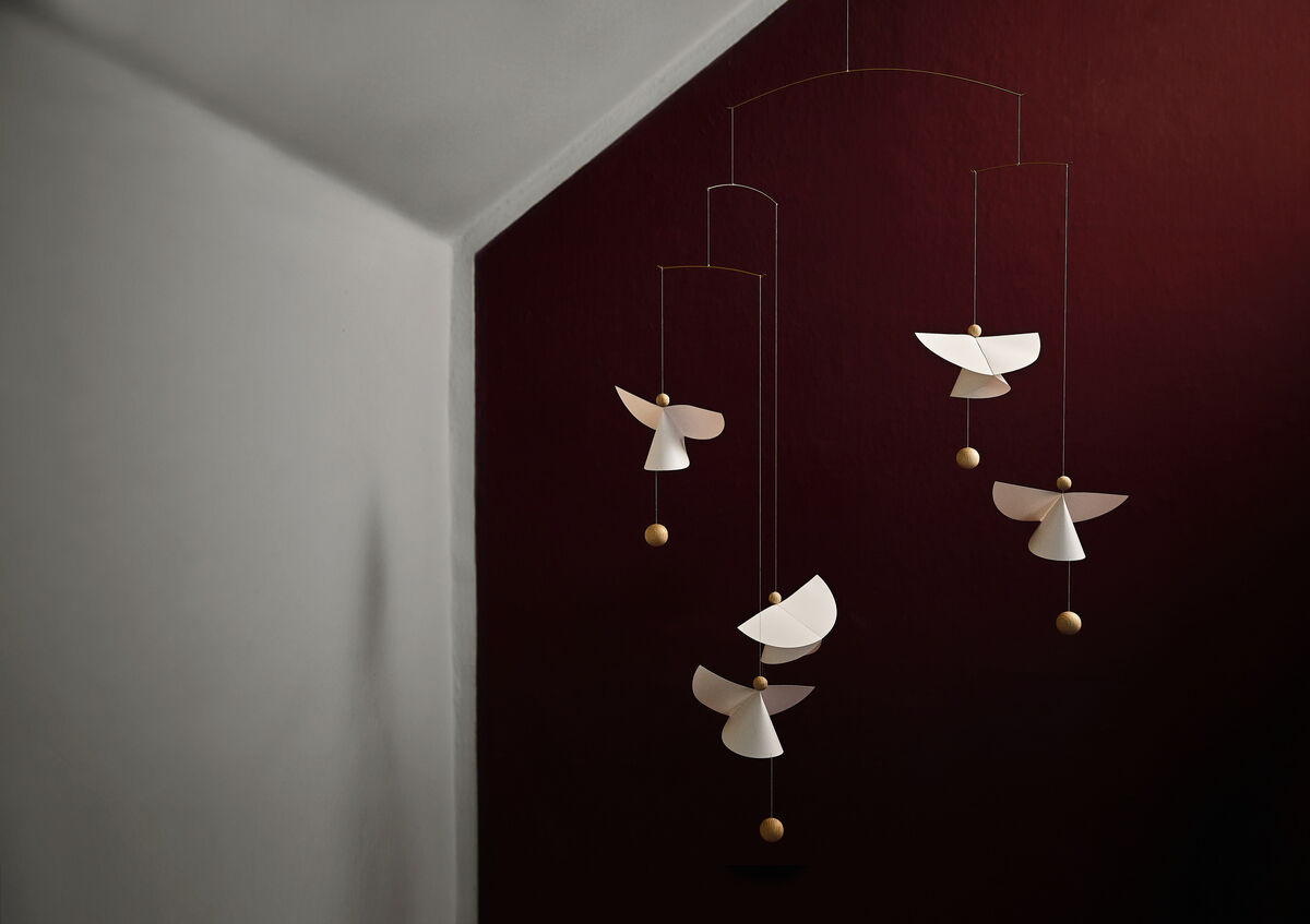 Mobile »Schutzengel 5« aus Holz mit fünf weißen Engel-Formen, die an dunkelroten Fäden hängen und einen minimalistischen Stil in einem Wohnraum unter schrägen Decken zeigt.