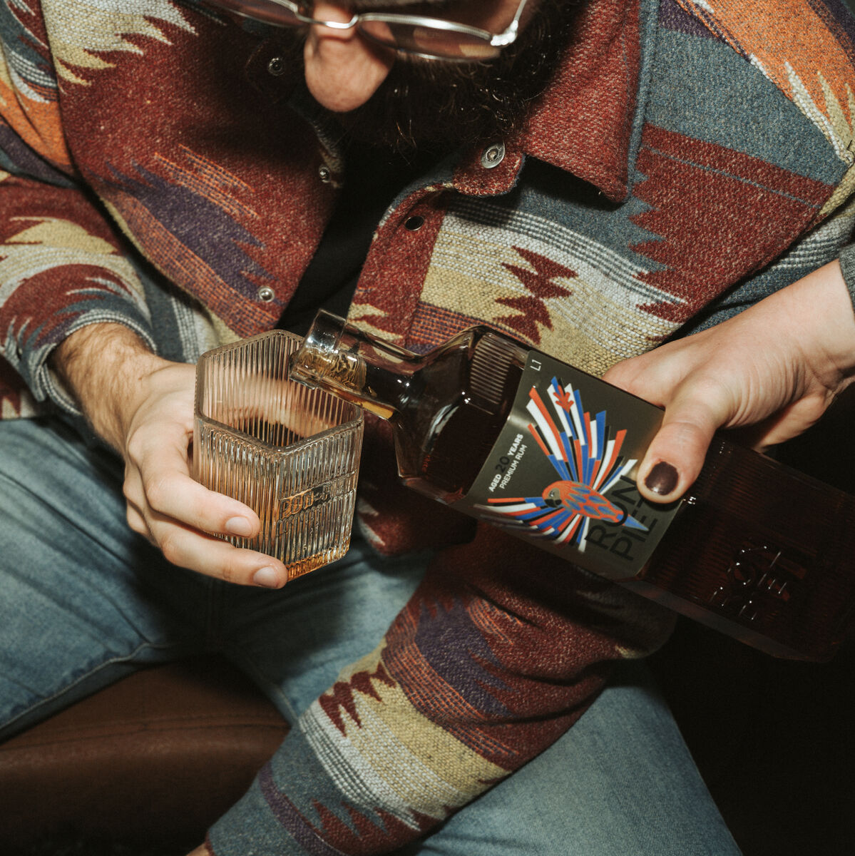 Eine Person in einer bunt gemusterten Jacke hält ein Glas, während eine andere Person eine Flasche Ron Piet Rum, 20 Jahre gereift, in das Glas einschenkt.