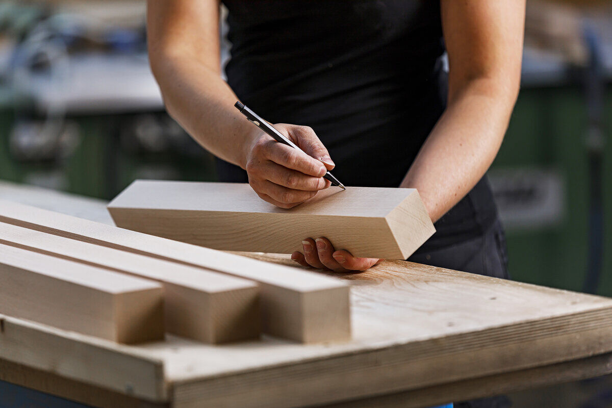 Eine Person arbeitet mit einem Holzblock auf einem Tisch. Der Holzblock wird mit einem Stift markiert. Weitere Holzblöcke sind auf dem Tisch zu sehen. Im Hintergrund sind unscharfe Werkzeuge und Werkbänke erkennbar.
