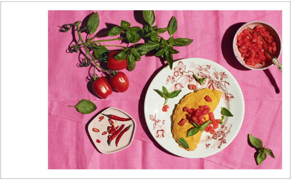 Ein Kochbuchbild mit einem schön angerichteten Omelett, garniert mit Tomatenwürfeln und Basilikumblättern. Daneben sind frische Tomaten und Kräuter, alles auf einem pinkfarbenen Tuch dargestellt.