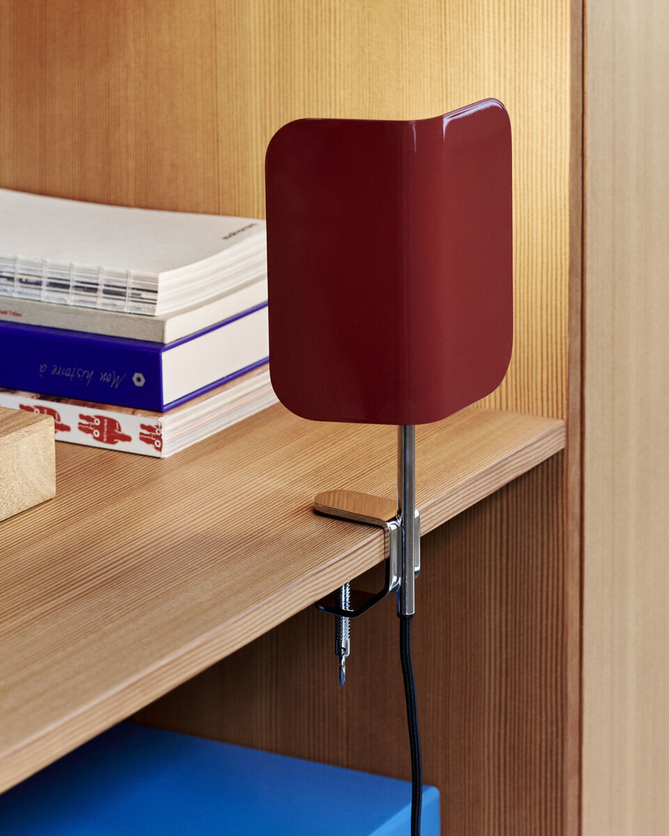 Rote Klemmlampe »Clip« von Apex an einem hölzernen Schreibtisch befestigt, mit zwei Schrauben und einem Kabel, umgeben von Büchern und anderen Gegenständen. Die Lampe hat ein minimalistisches, modernes Design mit einem silbernen Arm.