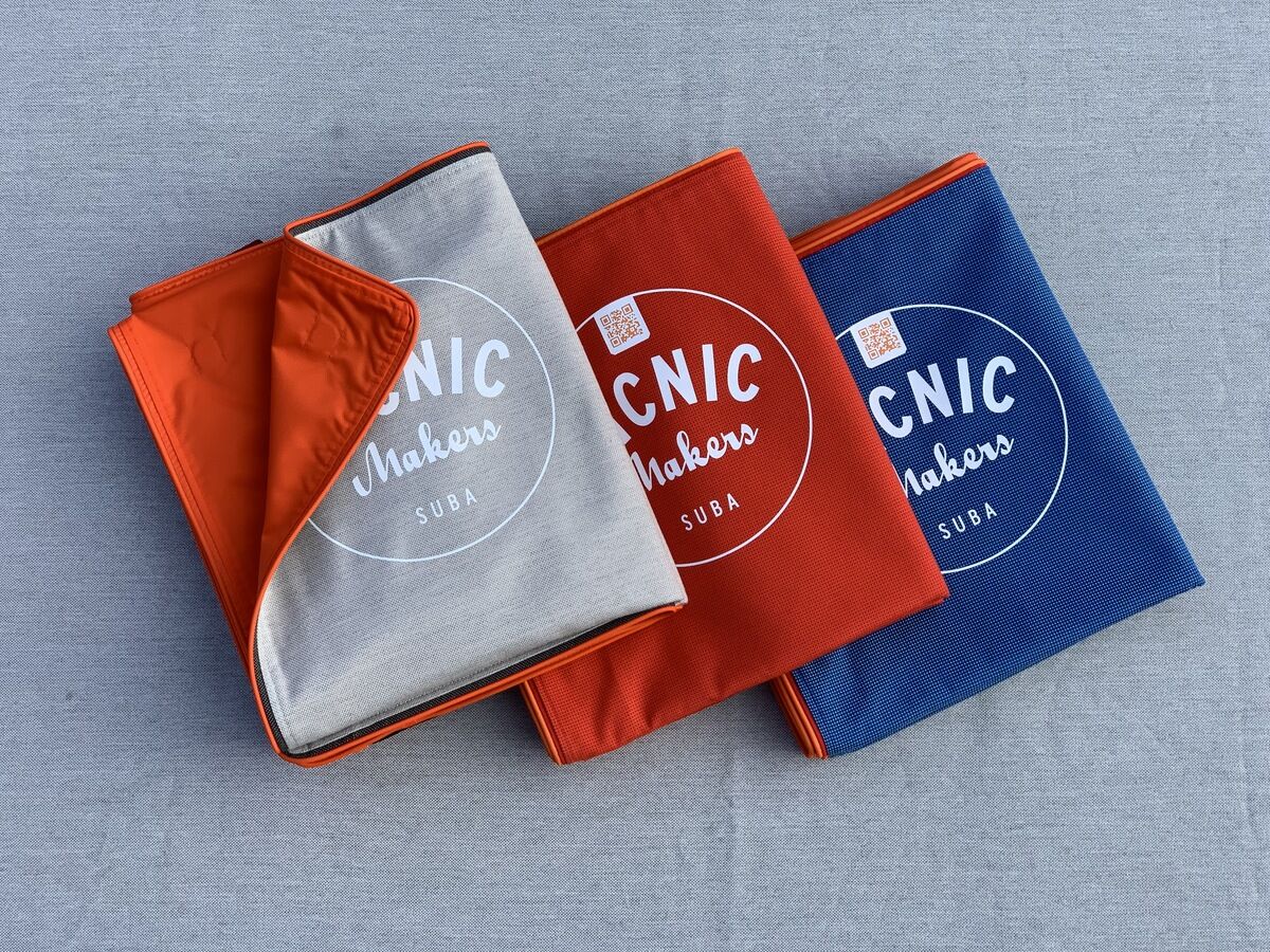 Drei zusammengefaltete Picknickdecken, jeweils in den Farben Grau, Orange und Blau, liegen nebeneinander auf einer grauen Oberfläche. Auf jeder Decke steht in weißer Schrift 'Picnic Makers Suba'.