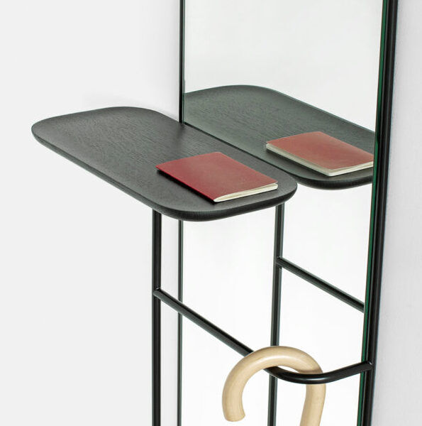 Ein moderner Wandspiegel der Marke »Concierge« mit einem kleinen Regal darunter. Auf dem Regal liegt ein rotes Notizbuch, und ein beiger Regenschirm mit blauem Griff hängt an einer Halterung unter dem Regal.