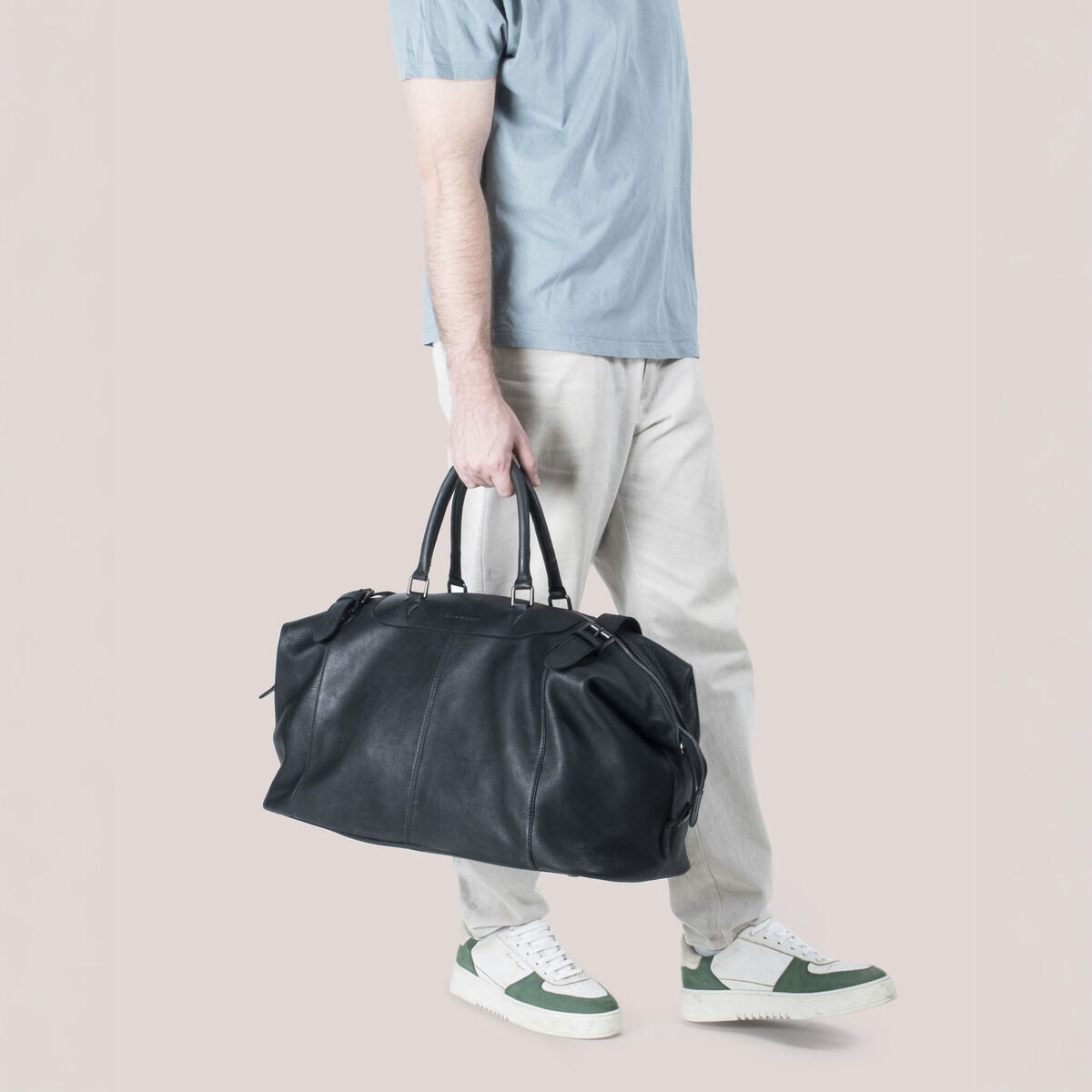 Ein Mann in heller Hose, blauen Sneakers und hellblauem T-Shirt trägt die schwarze ZEIT-Sonderedition Travelbag »Toro« von Harold's Lederwaren, gefertigt aus genarbtem, pflanzlich gegerbtem Rindleder. Die Tasche wirkt elegant, bietet viel Stauraum und besitzt einen abnehmbaren Tragegurt.