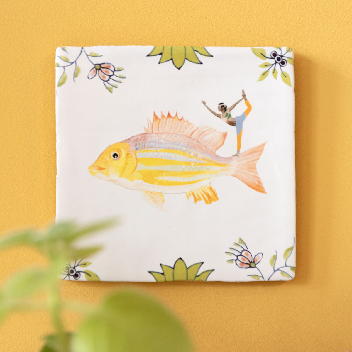 Keramikfliese mit einer handgemalten Darstellung eines gelben Fisches und grünem Blumenmuster an einer gelben Wand mit Pflanzen im Vordergrund.