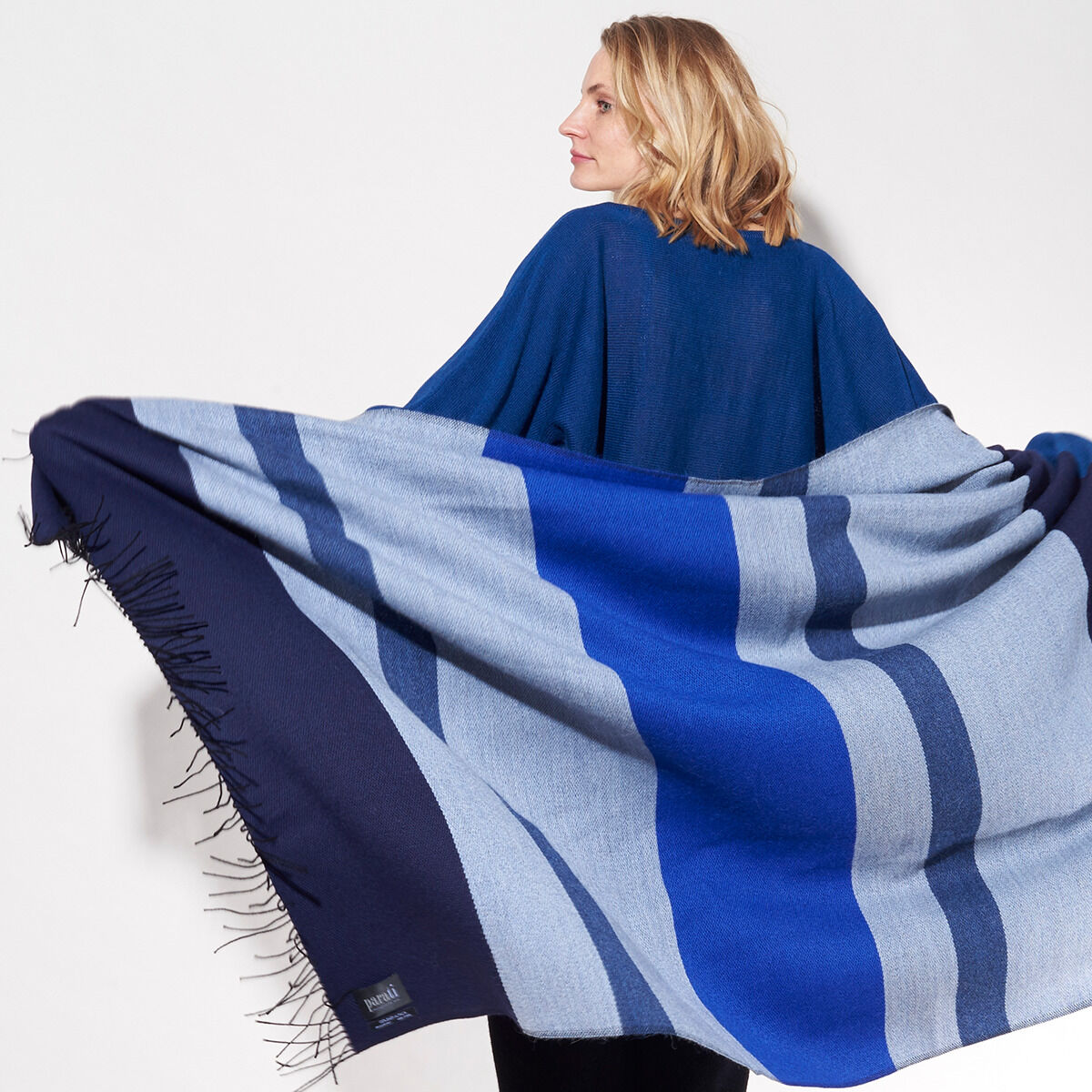Eine Frau trägt ein Cape der ZEIT-Jubiläumsedition »Shades of Blue«. Das Cape zeigt verschiedene Blau-Töne mit einem Streifenmuster und Fransen an den Kanten.