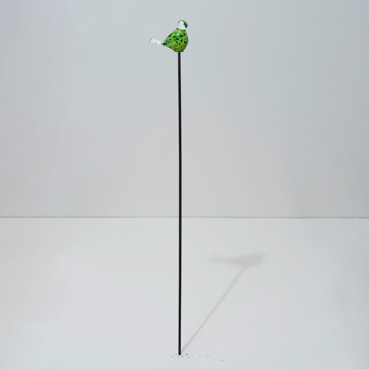 Der Gartenstecker Vogeltrio der Marke Ars mundi zeigt einen grünen Glasvogel mit weißen Akzenten auf einem sehr langen, dünnen Metallstab vor neutralem, lichtem Hintergrund. Die minimalistische Dekoration wirkt elegant und eignet sich ideal für moderne Gärten.