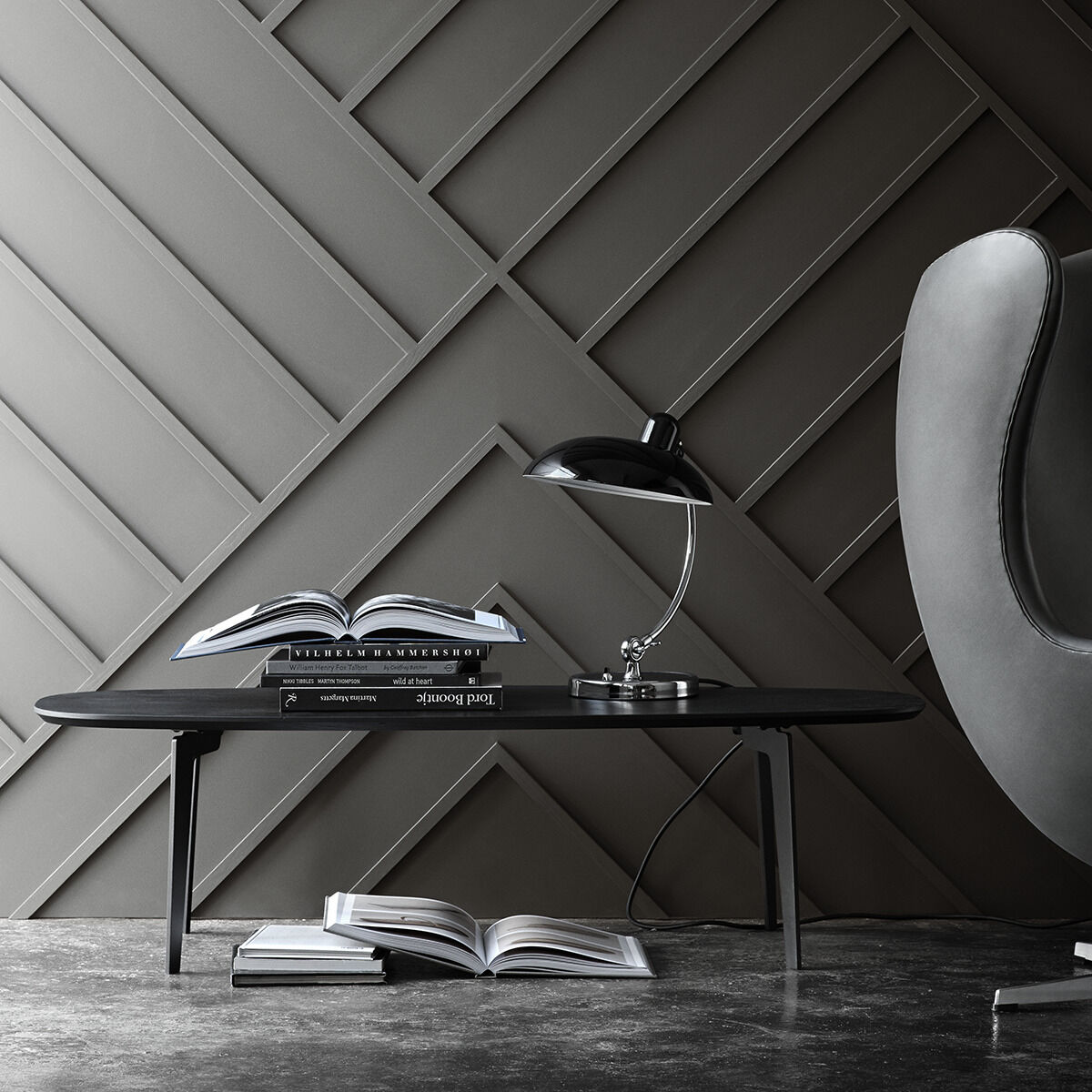 Eine schwarze Tischlampe des Modells »Kaiser Idell« steht auf einem modernen schwarzen Tisch, umgeben von aufgeschlagenen Büchern. Der Hintergrund zeigt eine graue, geometrische Wandgestaltung und einen modernen, stilvollen Stuhl.