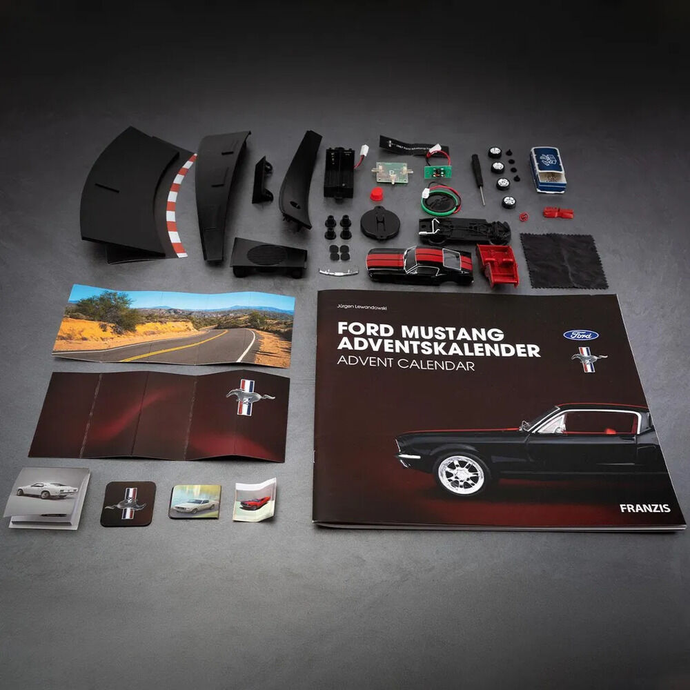 Auf dunklem Untergrund liegen die Teile des FRANZIS Ford-Mustang-Adventskalenders: schwarze Karosserieteile, rote und schwarze Modellkomponenten, Räder, Elektronik, Kabel, eine blaue Batteriebox und Stoff. Daneben liegen Begleitmaterialien und das Heft „Ford Mustang Adventskalender“ mit schwarzem Mustang Fastback.