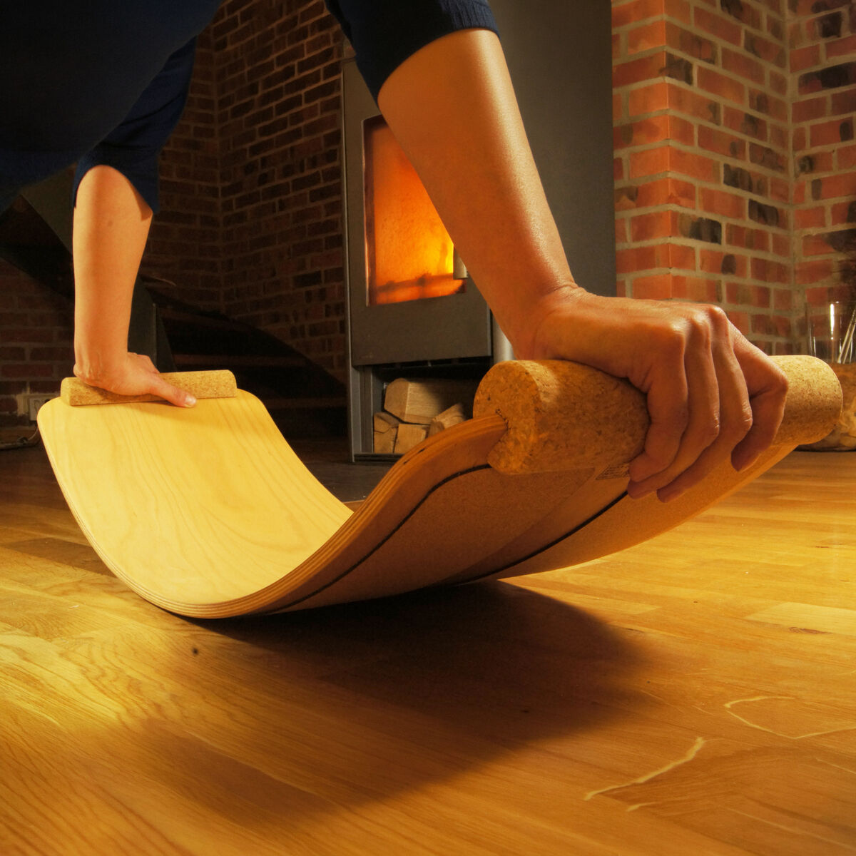 Person stützt sich mit beiden Händen auf ein Balance Board mit Korkrollen-Handgriffen von TicToys auf einem Holzfußboden ab. Im Hintergrund ist ein brennender Kamin vor einer Ziegelwand sichtbar. Das Bild vermittelt eine gemütliche Trainingsatmosphäre.