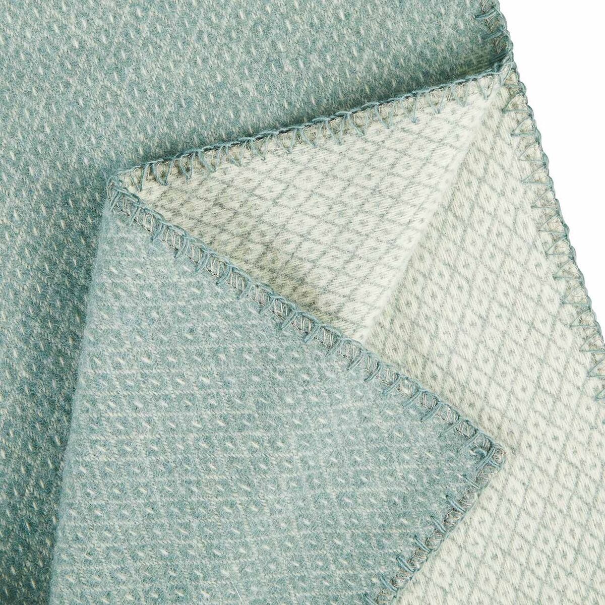 Eine zweifarbige Decke mit einer Seite in Blau und der anderen Seite in Cremeweiß, die in einer geometrischen Waffelstruktur gewebt ist. Die Kanten der Decke sind ordentlich vernäht.