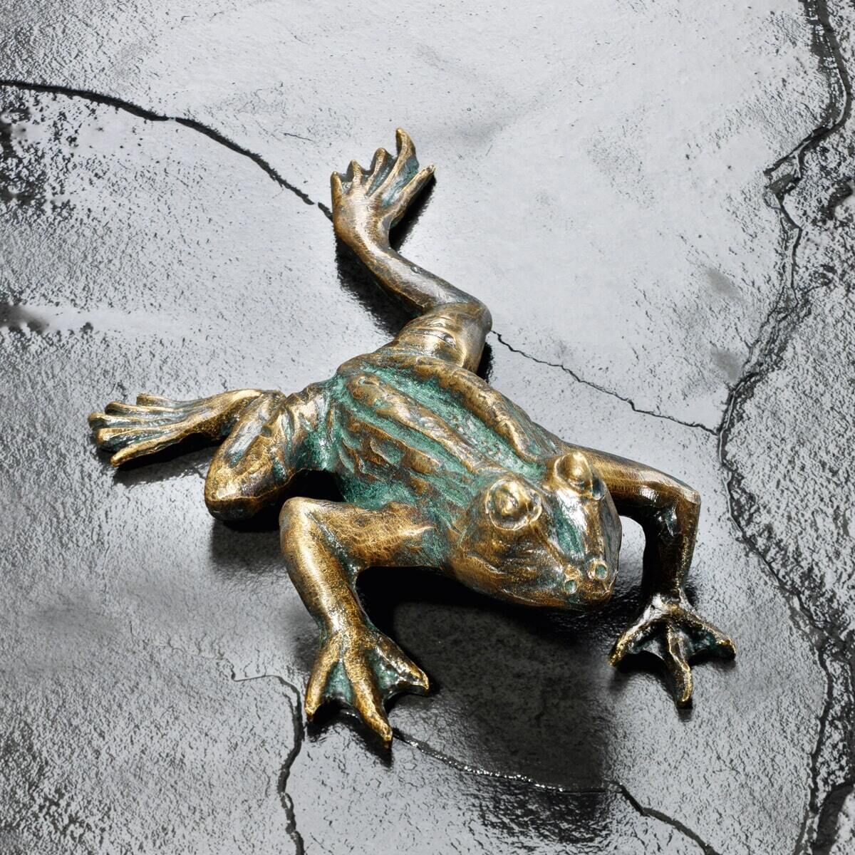 Gartenskulptur in Form eines Kletterfrosches, gefertigt aus Metall, in Bronzeoptik. Der Frosch hat detailliert ausgearbeiteten Körper und Arme, die eine kletternde Pose einnehmen. Die Skulptur befindet sich auf einem dunkelgrauen, strukturierten Hintergrund.