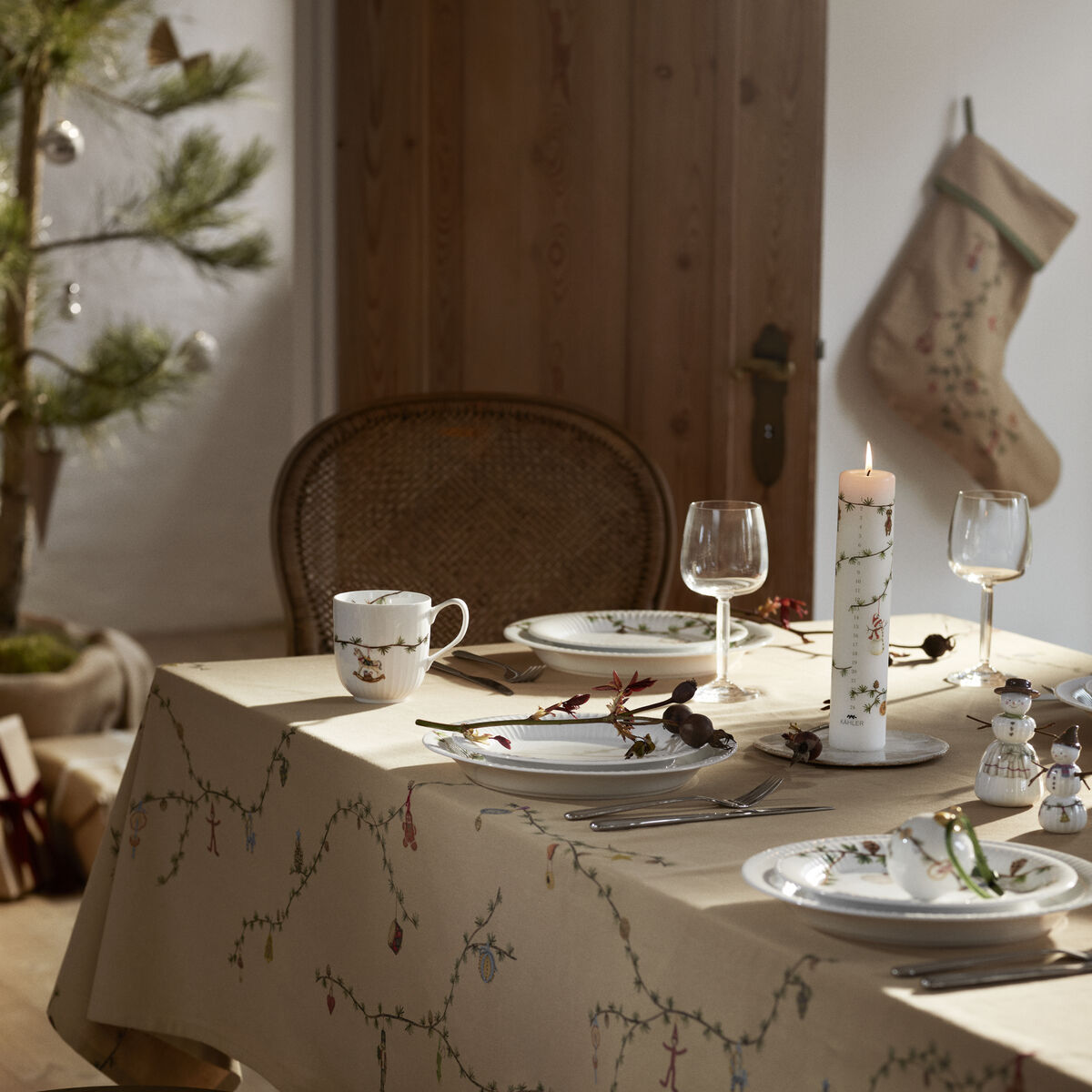 Beige Weihnachtsstrumpf der Marke Hammershøi hängt dekorativ an einer Holzwand in einem weihnachtlich dekorierten Zimmer. Im Vordergrund ist ein festlich gedeckter Tisch mit weiß-beiger Tischdecke und Weihnachtsgeschirr zu sehen. Die gemütliche Atmosphäre wird durch warmes Licht und dezente Weihnachtsdekoration unterstrichen.