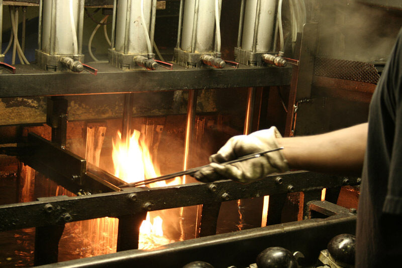 Eine Aufnahme einer industriellen Produktionsstätte, in der ein Messer geschmiedet wird. Eine Person mit Handschuhen hält einen heiß glühenden Metallstab über eine Feuerquelle, während Maschinen und Rohre im Hintergrund zu sehen sind. Strenge Sicherheitsvorkehrungen scheinen getroffen zu sein.