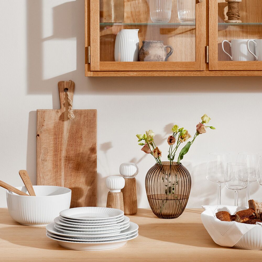 Auf einem hellen Holztisch steht eine stilvolle, weiße Gewürzmühle der Serie »Hammershøi«, umgeben von weißem Geschirr, Gläsern, einem Schneidebrett, einer Vase mit Blumen und Vitrinenschränken im Hintergrund. Die Szene zeigt ein modernes skandinavisches Küchendesign in ruhigen Natur- und Weißtönen.