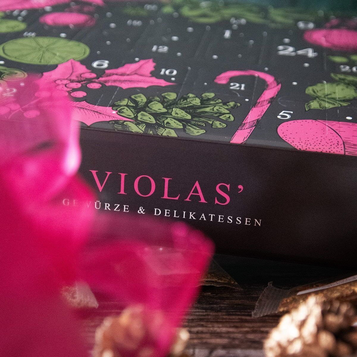 Ein Adventskalender von Violas, verziert mit grünen Blättern und rosa Kugeln. Der Titel 'VIOLAS' Gewürze & Delikatessen' ist auf der Vorderseite zu sehen. Im Vordergrund sind unscharfe Rosa-Texturen erkennbar.