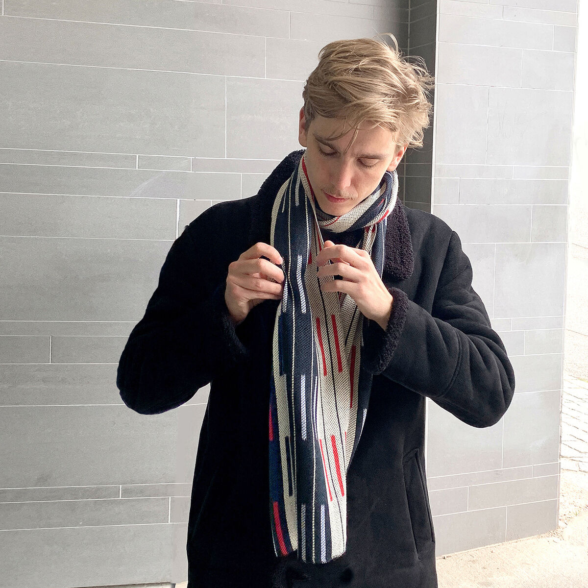 Ein Mann mit blondem Haar trägt einen Seidenschal »Doublé« in Grau mit roten und weißen Akzenten. Er trägt außerdem einen schwarzen Mantel und steht vor einer grauen Wand.