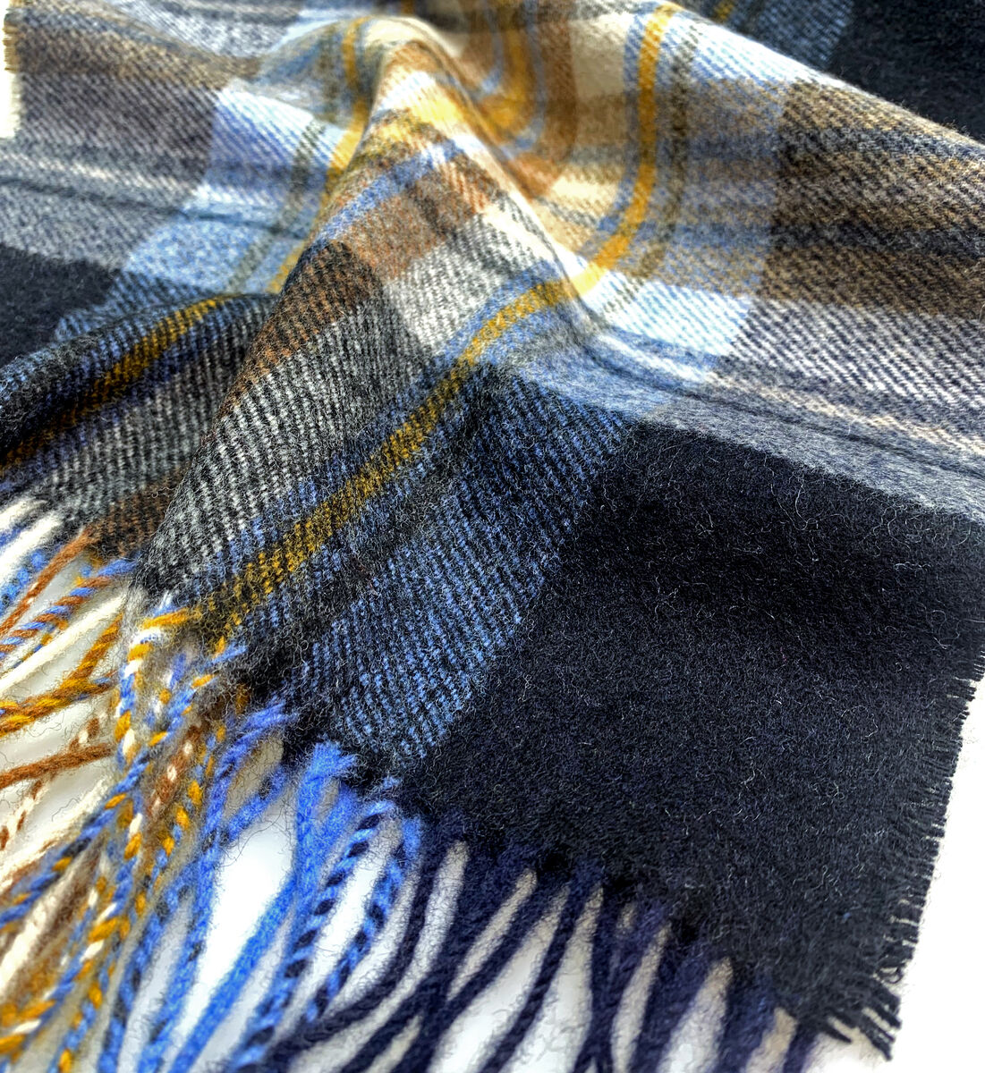 Nahaufnahme eines hochwertigen Kaschmirschals mit Karomuster in Blau-, Schwarz-, Braun-, Weiß- und Gelbtönen. Die Fransen des Schals liegen locker übereinander, das Material wirkt weich und elegant.