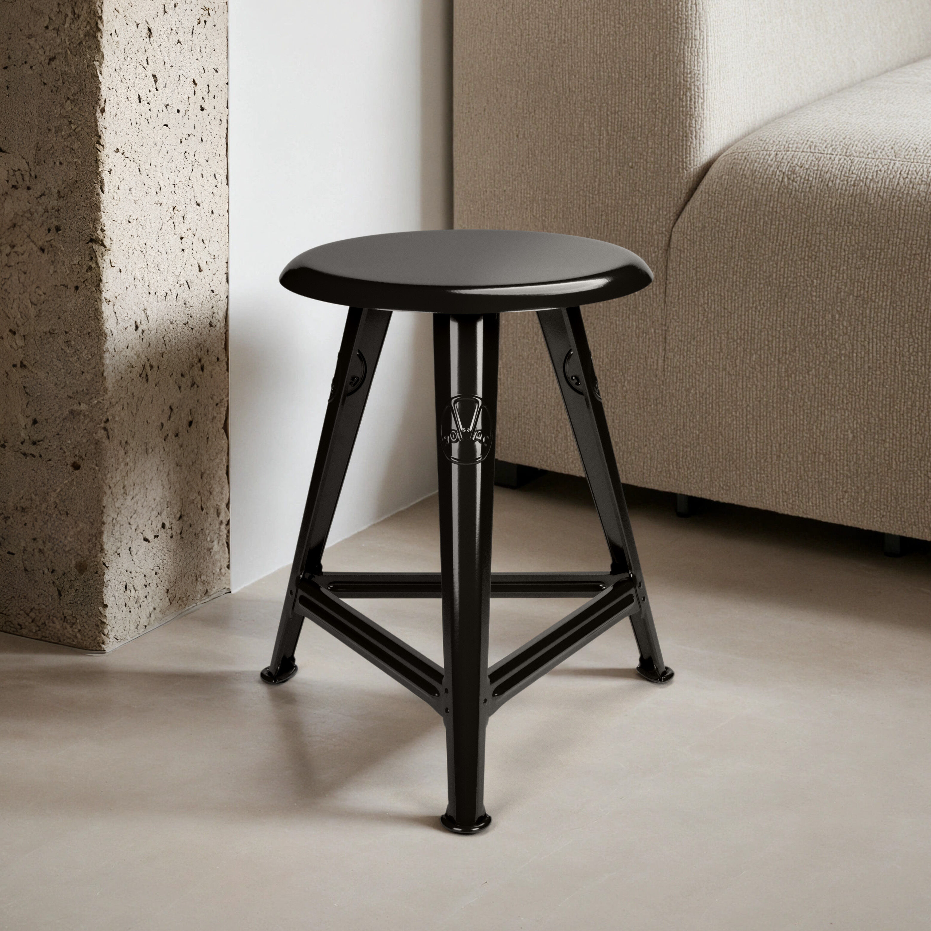Der schwarze Rowac-Schemel »Just Steel« von ROWAC steht auf einem hellgrauen Boden in einem modernen, hellen Innenraum vor einer gemauerten Wand und neben einem hellbeigen Stoffsofa. Der Hocker besteht aus glänzend pulverbeschichtetem, kaltgewalztem Stahl und wirkt stabil sowie elegant.